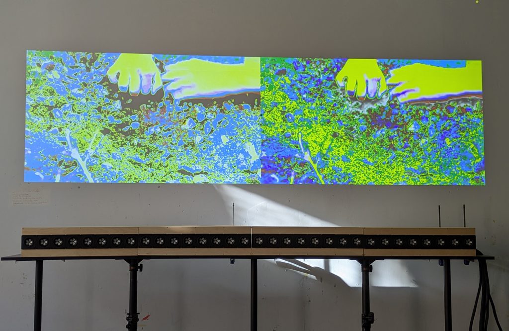 A photo of a 2 channel video, vividly colored in blue and green, of hands digging in soil. Below the video is a wavefield synthesis speaker array.